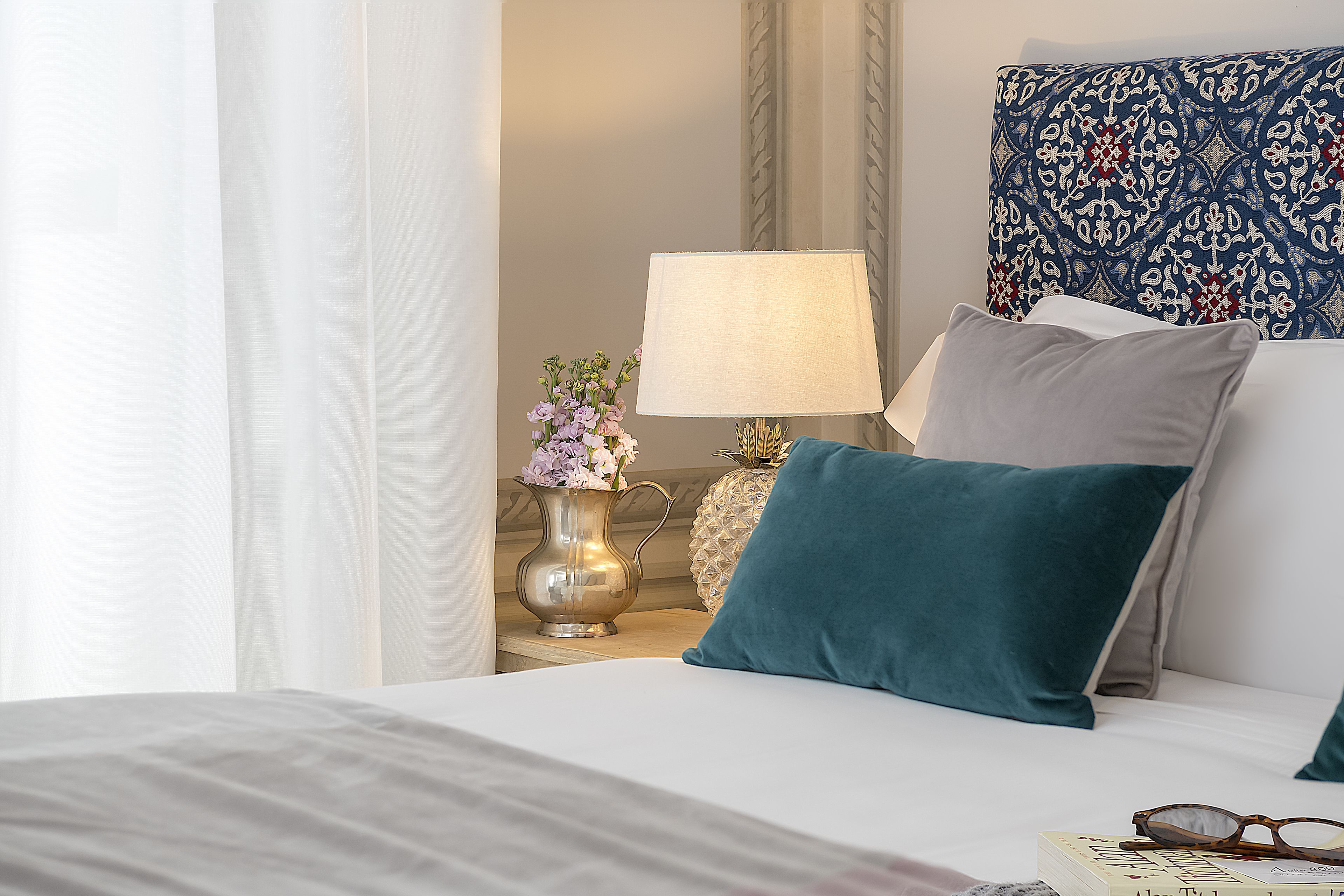 Un letto rifatto con un copriletto bianco e cuscini color petrolio, una lampada da comodino e un vaso con fiori su un tavolino.