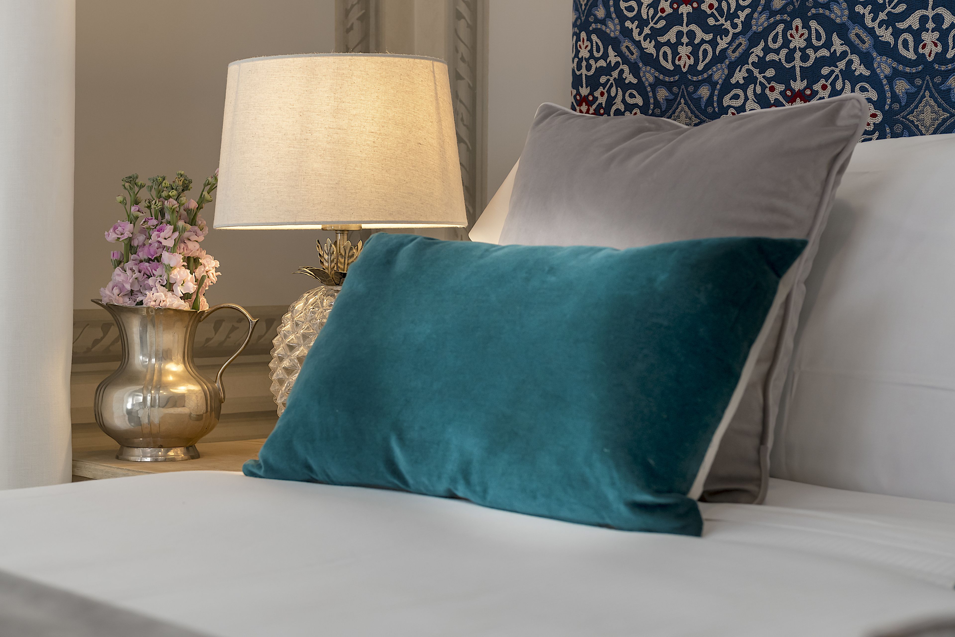 A bed with a teal pillow, a white pillow, a lamp, and a vase with flowers.