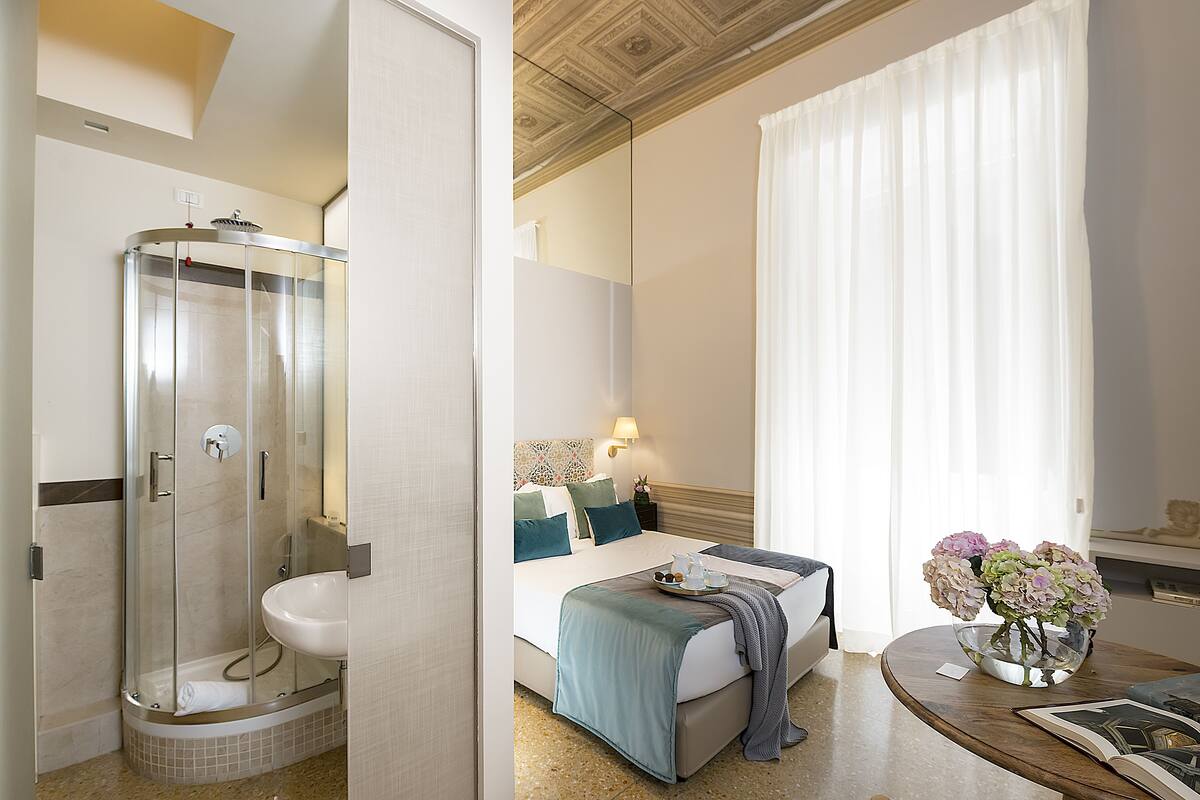 Habitación de hotel con ducha, cama con vistas, mesita de noche con un jarrón de flores y un amplio ventanal con cortinas.