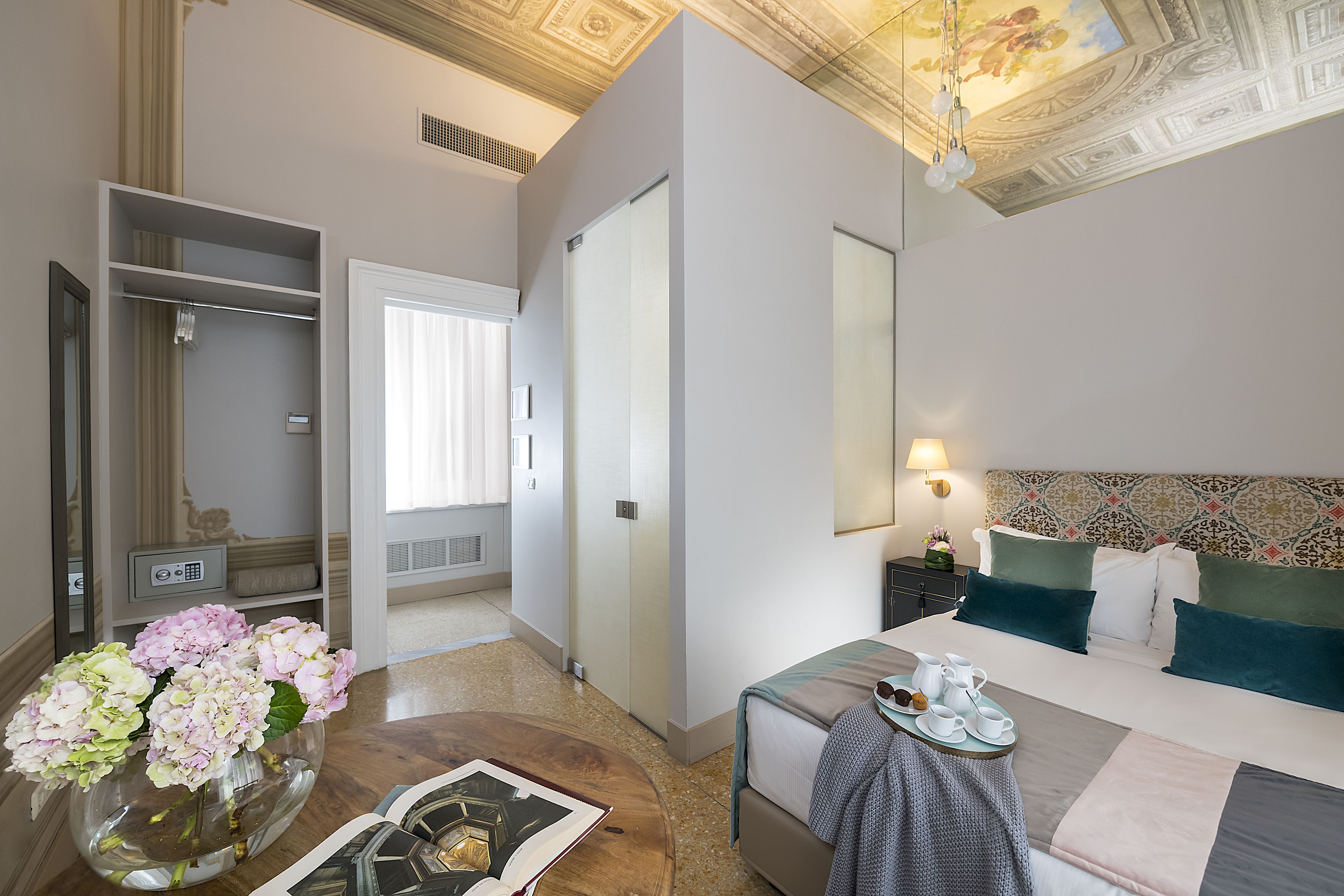Habitación de hotel con cama, mesita de noche y un espejo grande. Se observa un armario con perchero y una ventana con cortinas.