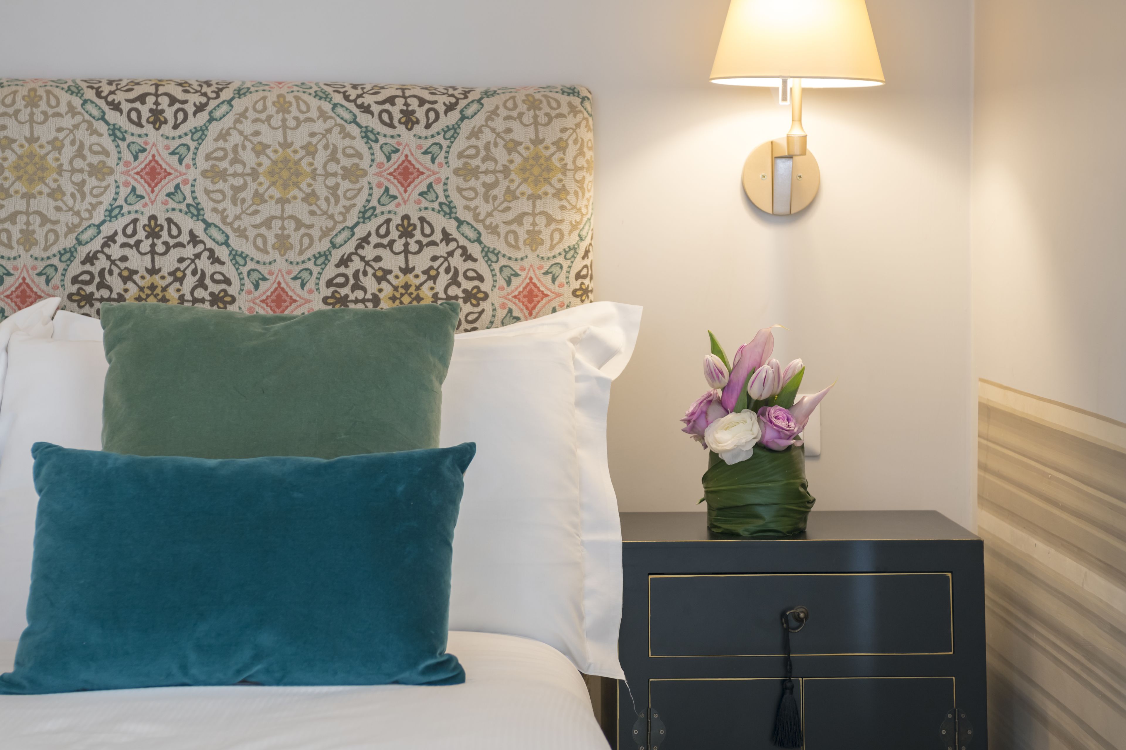 A neatly made bed with teal and white pillows, a patterned headboard, a bedside table with a vase of flowers, and a wall-mounted lamp.
