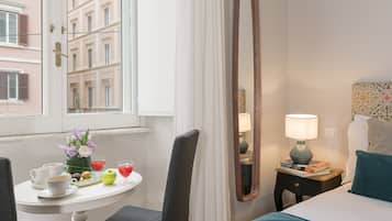 Ein Hotelzimmer mit einem Bett, einem Nachttisch mit Lampe, einem Sessel, einem Fenster mit Vorhängen und Blick auf Gebäude.