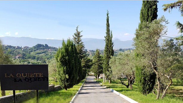 Un letrero que dice 'LA QUIETE LA QUIET' sobre un camino pavimentado bordeado de árboles, que conduce hacia un paisaje montañoso.