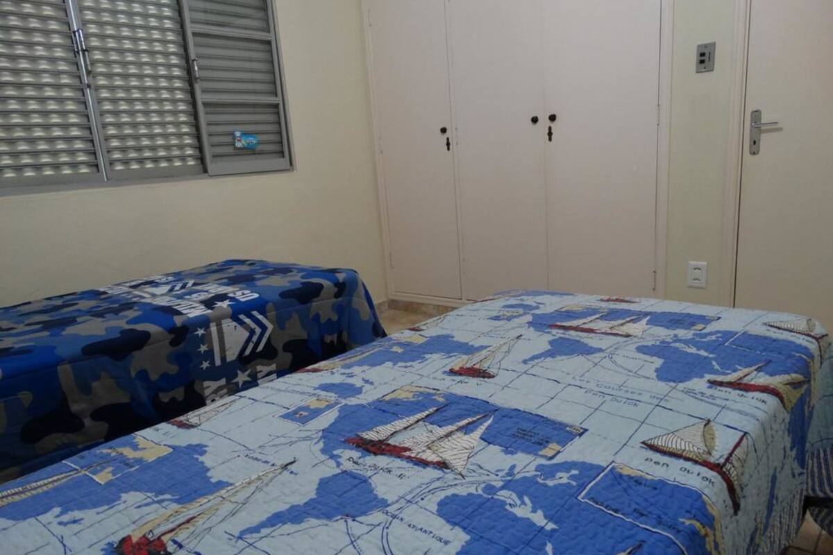 A bedroom with two beds, one with a blue camouflage bedspread and the other with a nautical-themed bedspread. There is a wardrobe with two doors and a window with blinds.