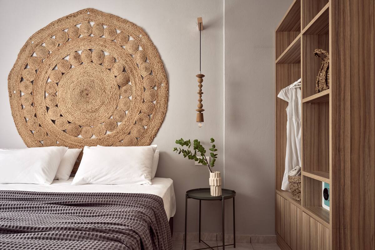 A bedroom with a large woven headboard, a bed with white linens, a small side table with a plant, and a wooden wardrobe.
