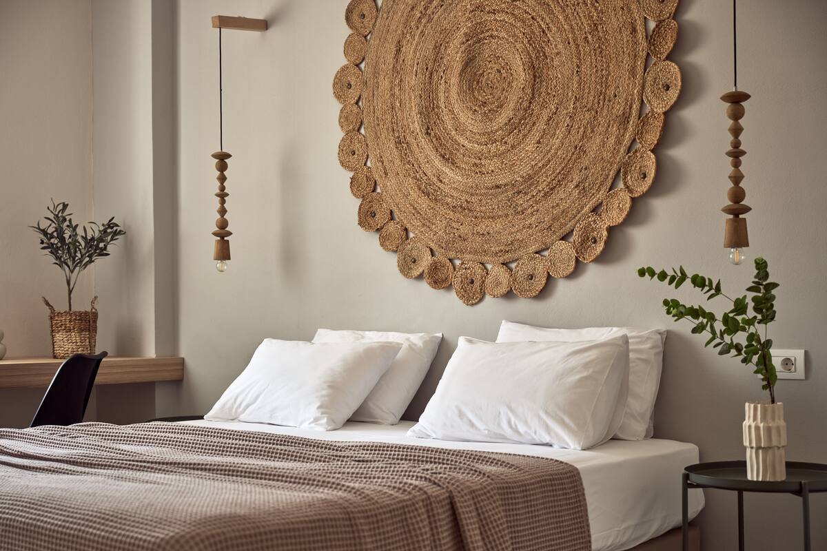 A bedroom with a large woven wall hanging, a bed with white linens, a checkered throw blanket, and a small side table with a plant.