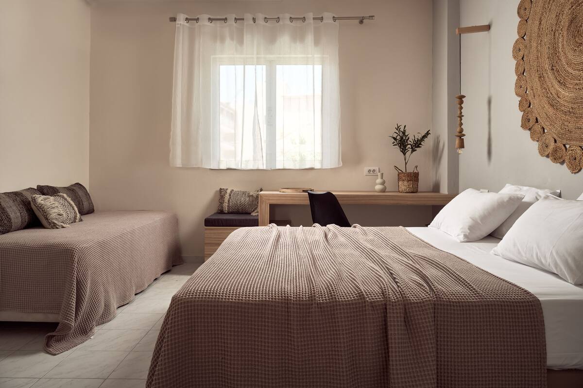 A bedroom with two beds, a window with sheer curtains, a bedside table with a lamp, and a woven wall hanging.