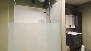Uma casa de banho moderna com divisória de vidro, chuveiro com cabeça de chuva e lavatório branco, além de um armário escuro.