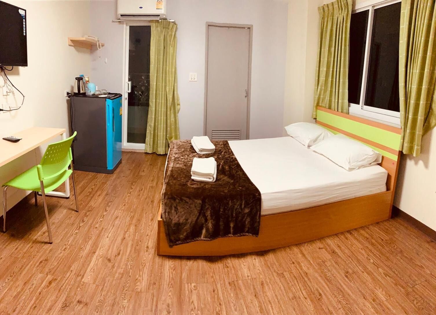 A hotel room with a wooden floor, a bed with white linens, a desk with a chair, a small kitchenette, and a window with curtains.