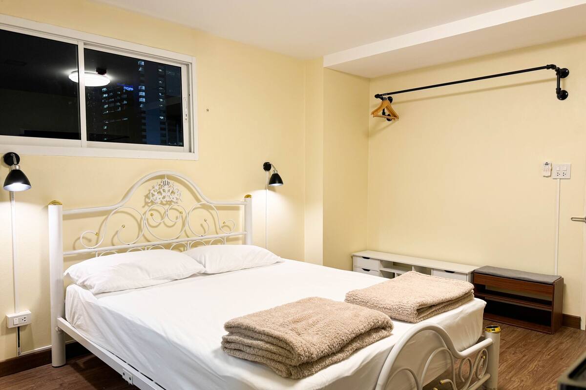 A neatly made bed with white linens and two beige towels, a bedside table with a lamp, and a window with a view of a cityscape at night.