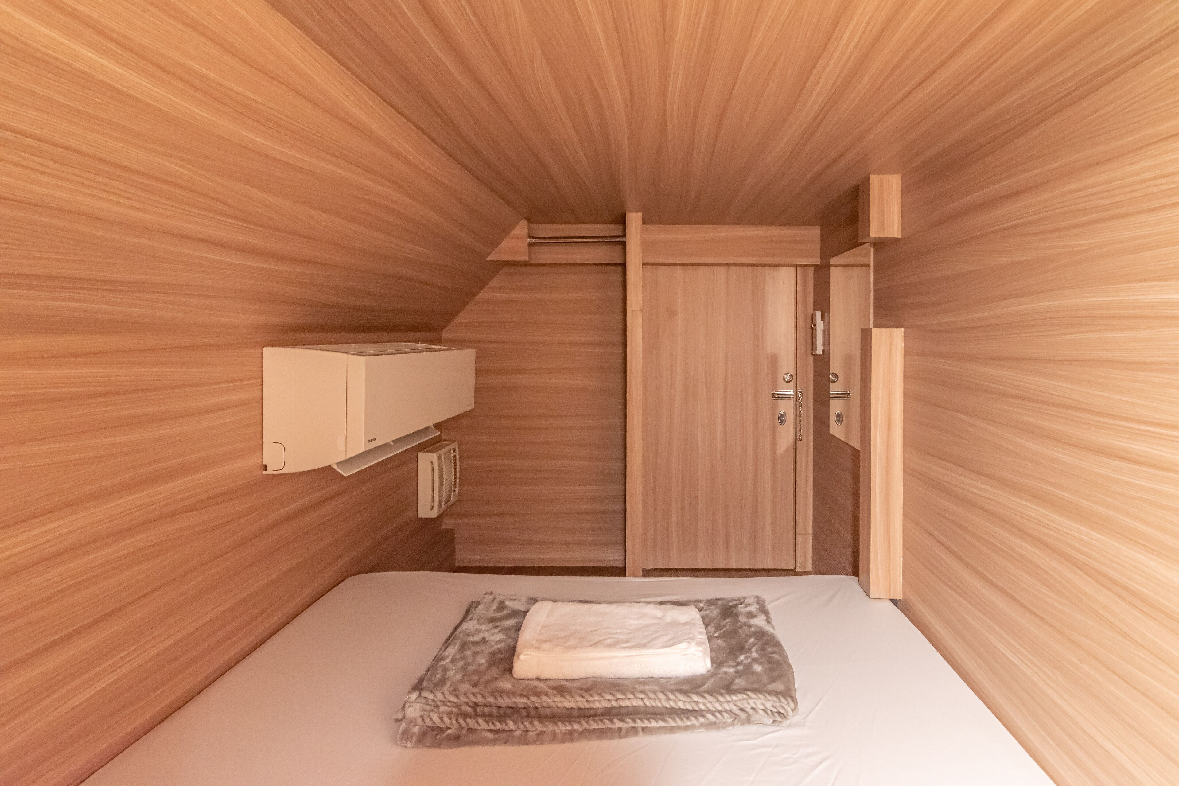 A compact bedroom with a wooden sloped ceiling, a bed with a white mattress and a gray blanket, a wooden wardrobe, and a small bedside table with a lamp.
