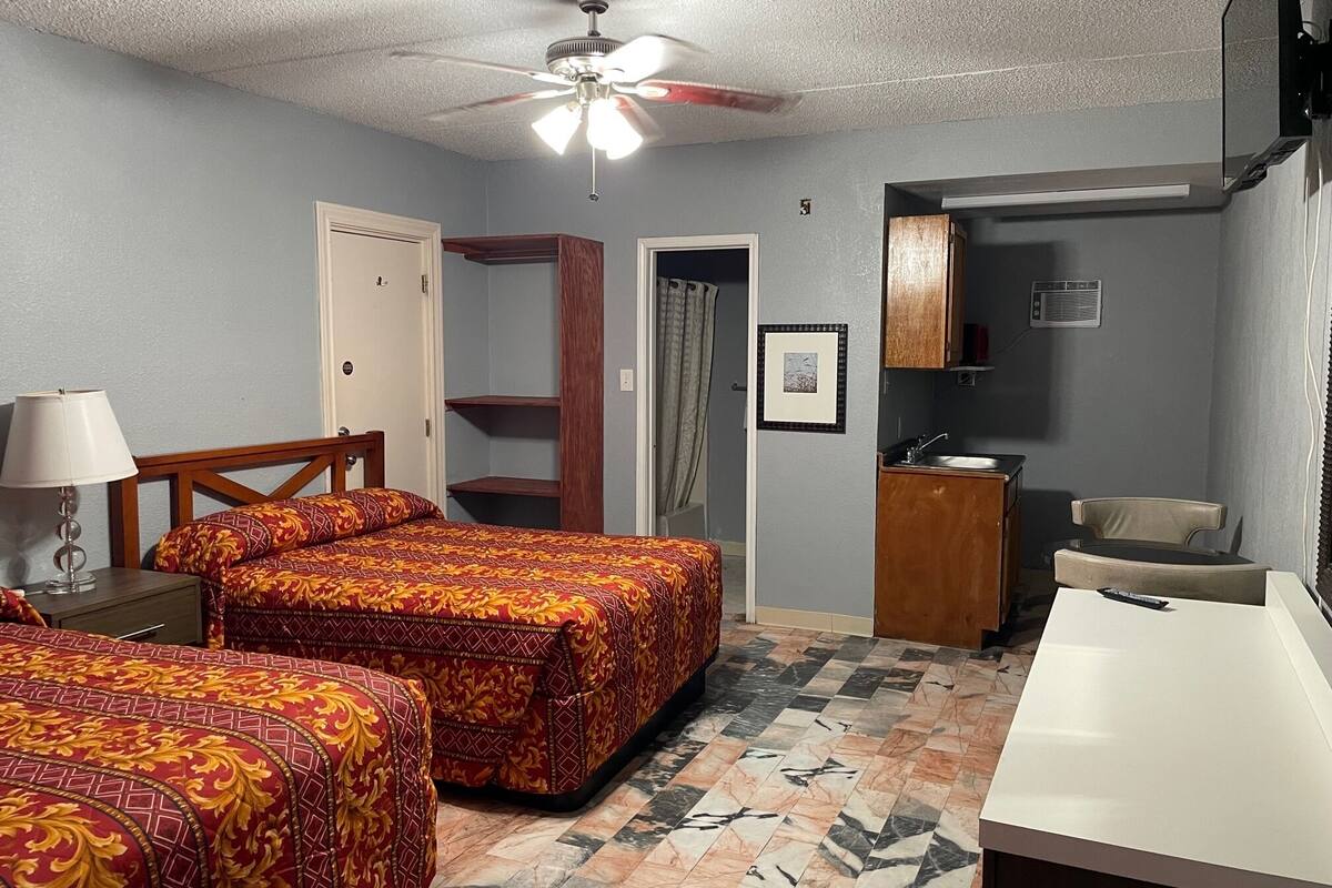 A hotel room with two beds, a desk, a chair, a ceiling fan, and a bathroom visible through an open door.