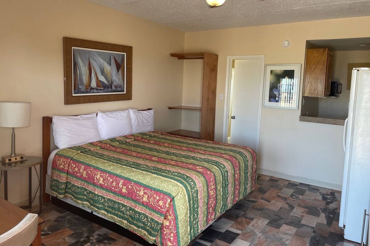 A hotel room with a bed, a ceiling fan, a painting, a door, and a kitchenette.