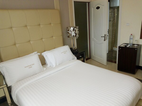 A hotel room with a large bed, two pillows, a bedside table with a lamp, and a door leading to a bathroom. - Hotel Amaritsah (Brazzaville)