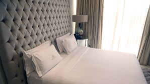 A neatly made bed with a tufted headboard, a bedside table with a lamp, and a window with curtains. - Hotel Amaritsah (Brazzaville)