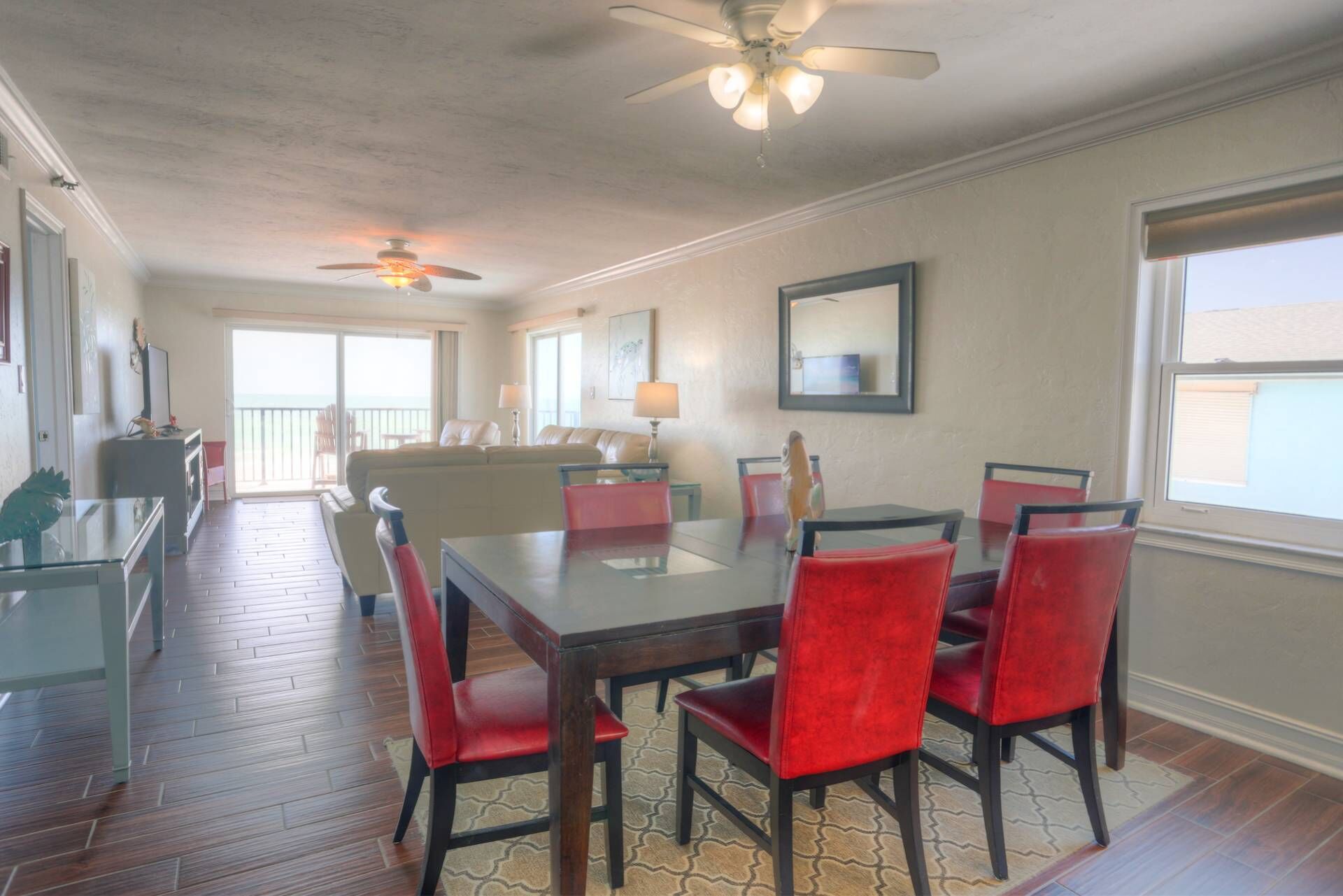 A dining area with a glass table and red chairs, a ceiling fan, and a view of the outdoors.