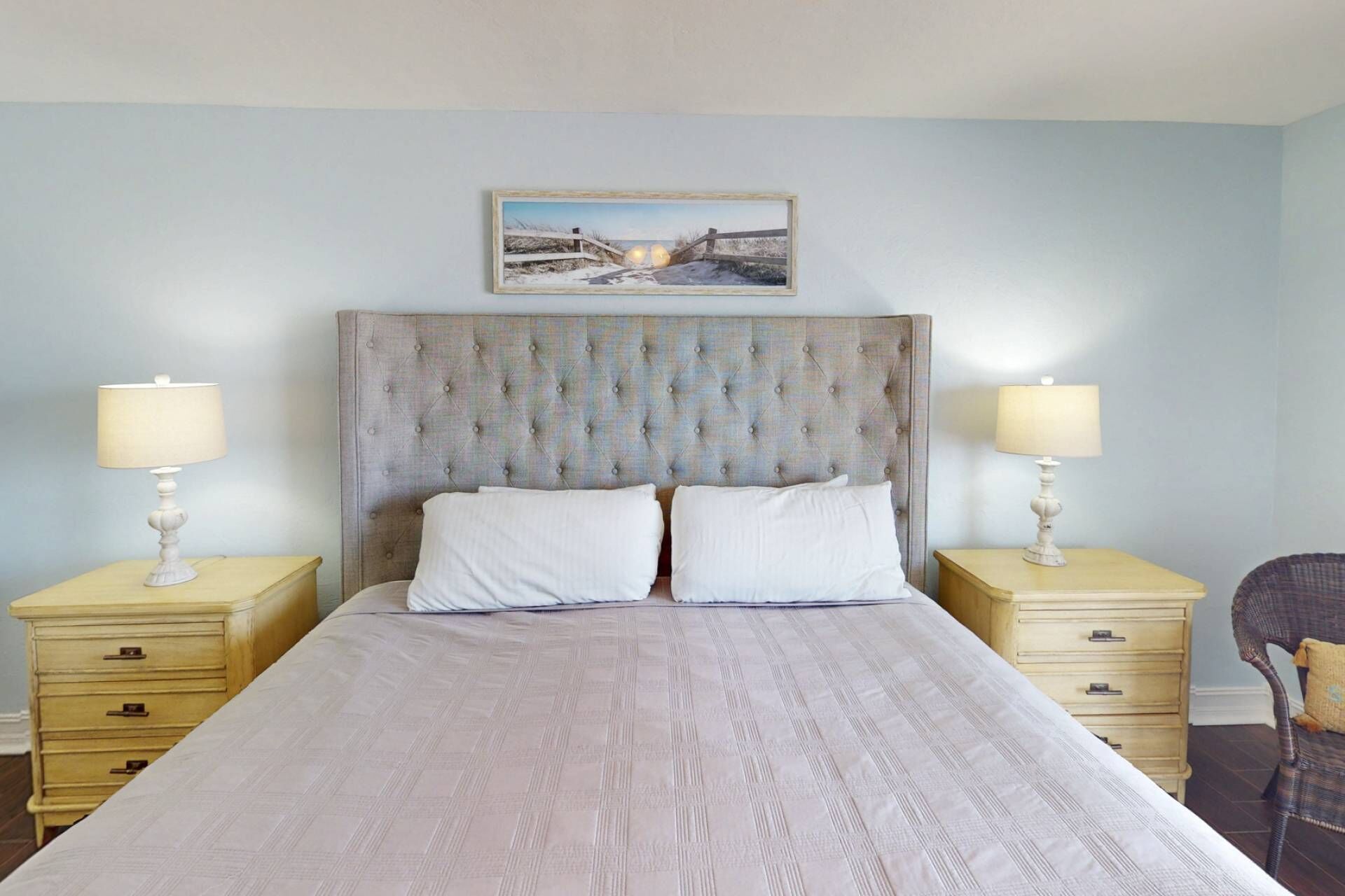 A bedroom with a tufted headboard, two bedside tables with lamps, a bed with white linens, and a framed picture above the headboard.