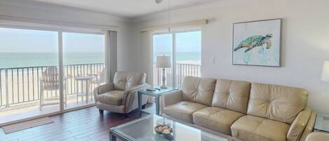 A spacious living room with a glass coffee table, a beige sofa, and a chair. There is a ceiling fan and a large window overlooking the ocean.