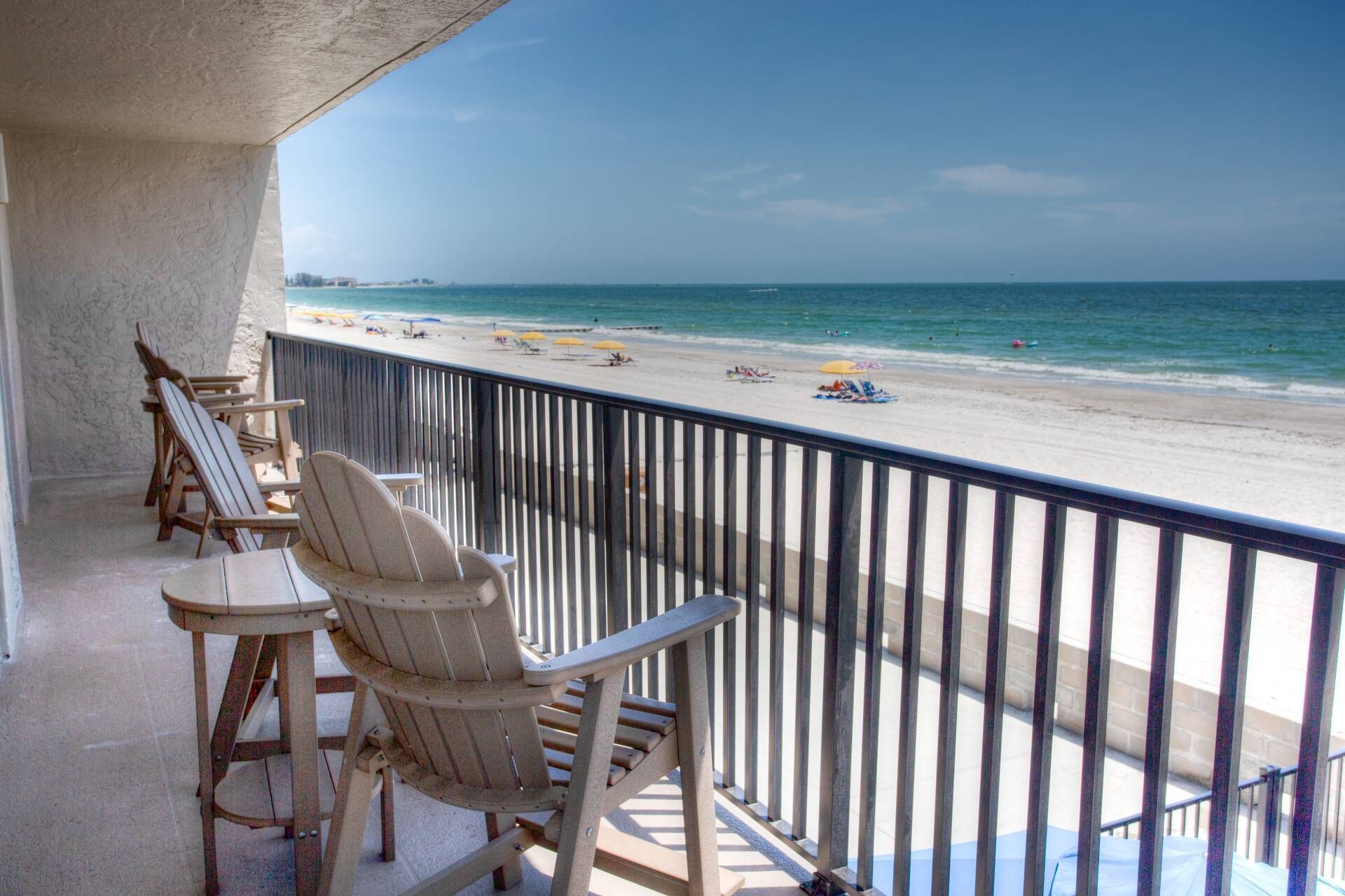 A balcony with Adirondack chairs overlooking a beach with umbrellas and people.