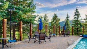 An outdoor pool area with chairs and umbrellas, surrounded by a wooden fence and trees. - Mtn Views-Heated Pool-HotTub-Grill-Deck+Walk to Mtn/Trails/Resort/Eat/Drink/Free Town Bus-Immaculate (Steamboat Springs)