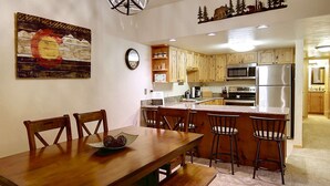 A modern kitchen with wooden cabinets, a stainless steel refrigerator, and a dining area with a wooden table and chairs. - Mtn Views-Heated Pool-HotTub-Grill-Deck+Walk to Mtn/Trails/Resort/Eat/Drink/Free Town Bus-Immaculate (Steamboat Springs)