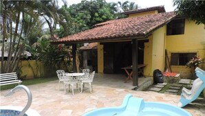 A swimming pool with a stone deck, a covered patio with a table and chairs, and a yellow building with a tiled roof.