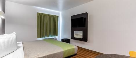 A modern hotel room with a bed, a small table, a chair, a flat-screen TV mounted on the wall, and a window with curtains.