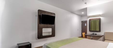 Una habitación de hotel con una cama, un televisor de pantalla plana montado en la pared, una mesita con una silla, un espejo y un calentador en el suelo.