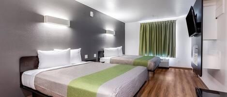 A hotel room with two beds, a flat-screen TV, a window with green curtains, and a wooden floor.