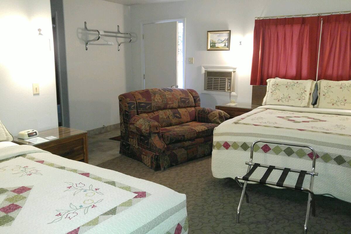 A bedroom with two beds, a patterned armchair, a bedside table, a wall-mounted heater, and a window with red curtains.