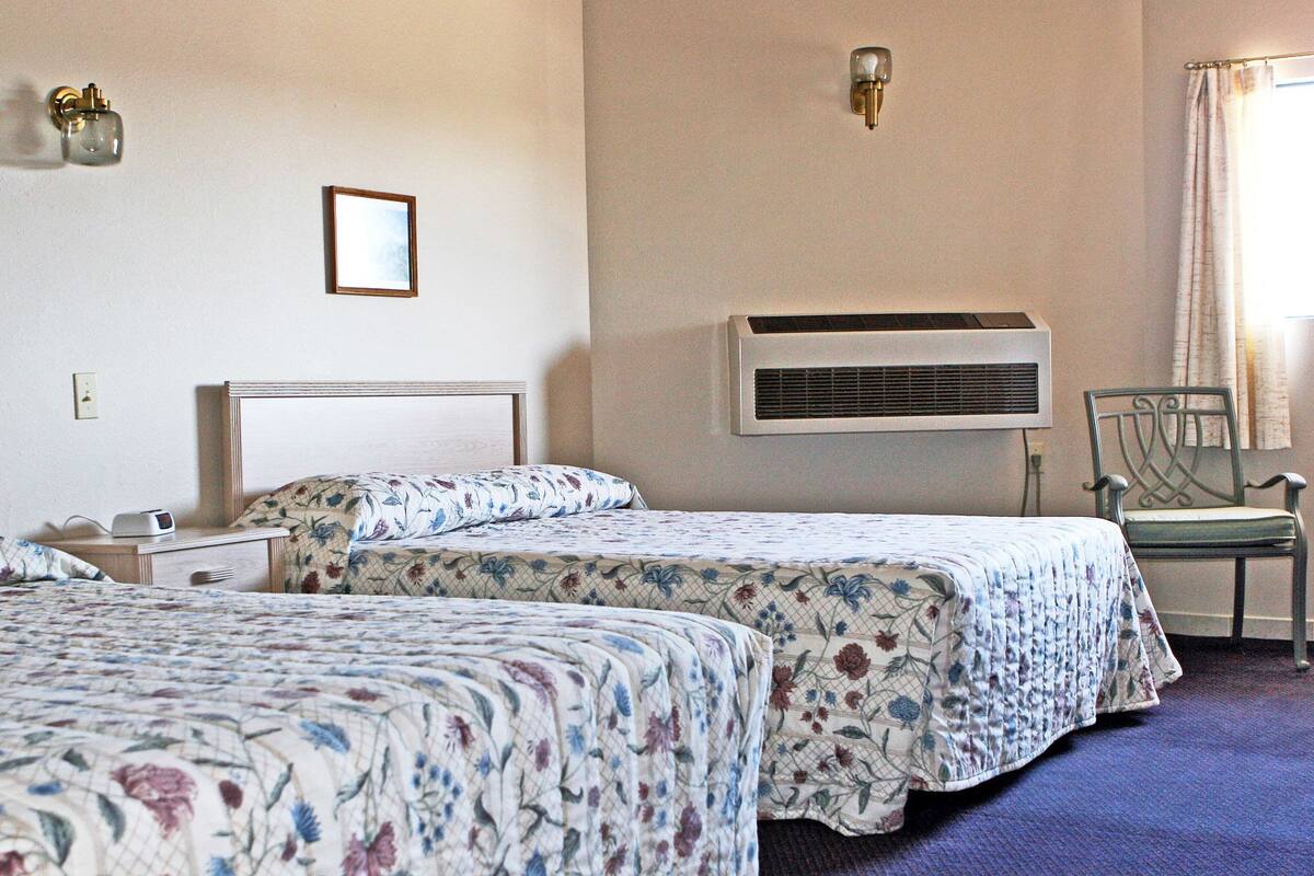 A hotel room with two beds, a chair, a nightstand, a wall-mounted air conditioner, and a picture on the wall.