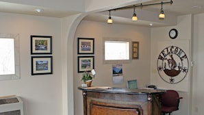 A reception area with a wooden reception desk, framed pictures on the wall, a welcome sign, and a clock. - Carter Mountain Motel (Cody)