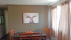 A dining area with a table and four chairs, a fruit basket on the table, and a painting of a deer on the wall. - Carter Mountain Motel (Cody)