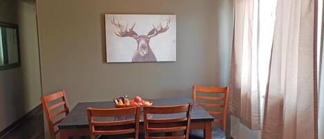 Uma sala de jantar com uma mesa e quatro cadeiras, uma cesta de frutas na mesa e um quadro de um veado na parede.