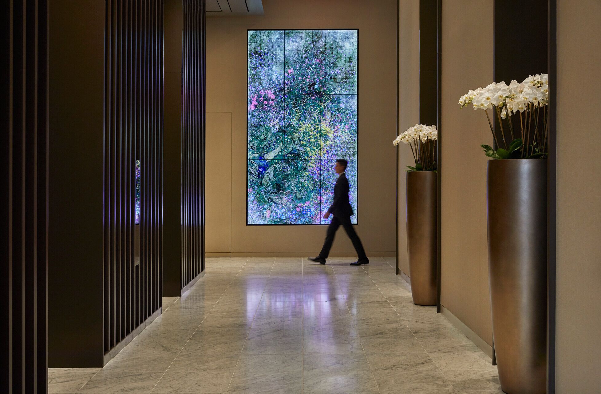 A corridor with a large digital art display, marble flooring, and tall planters with white flowers.