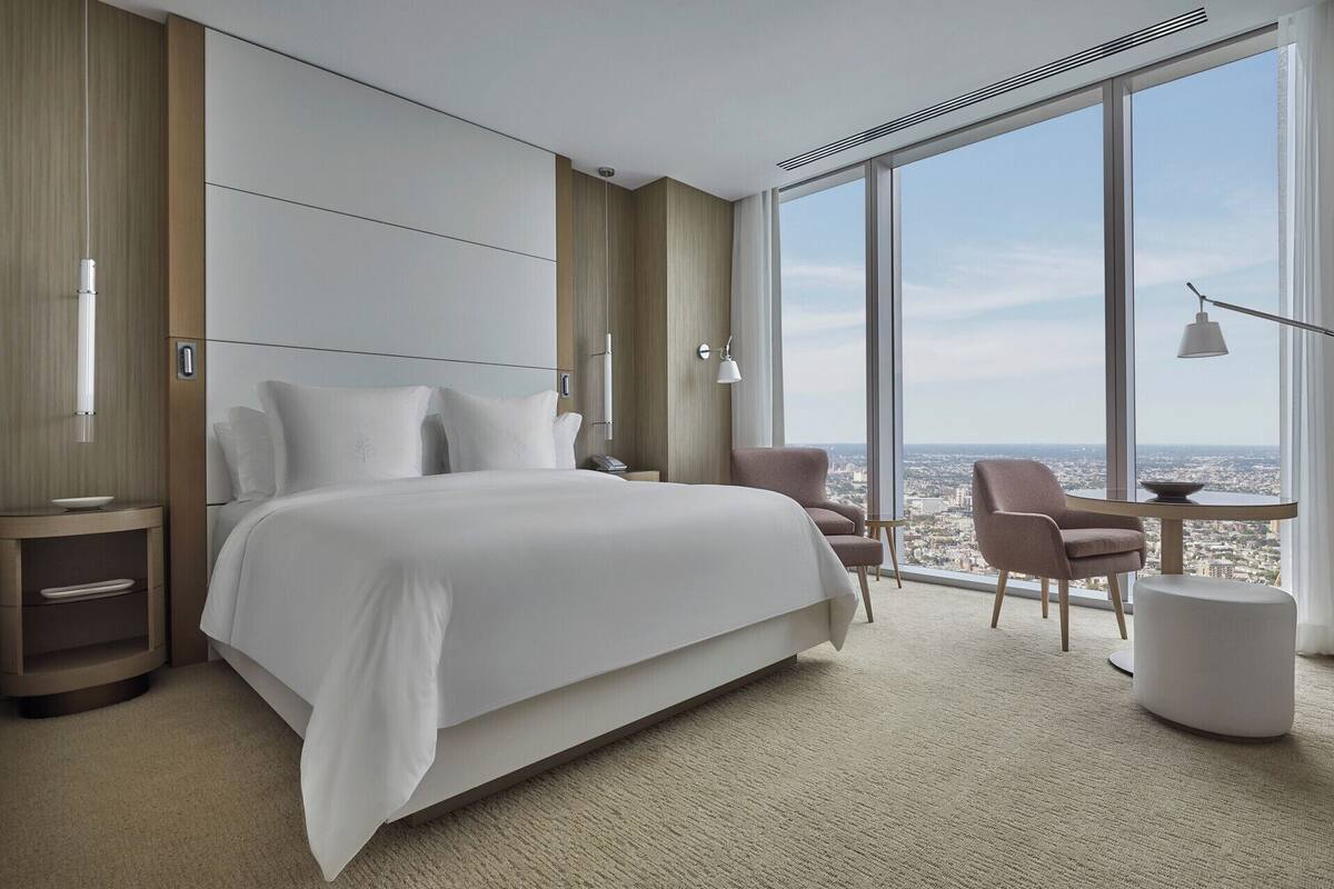 Ein modernes Hotelzimmer mit einem großen Bett, zwei Nachttischen, einem Sessel und Blick auf die Stadt durch große Fenster.