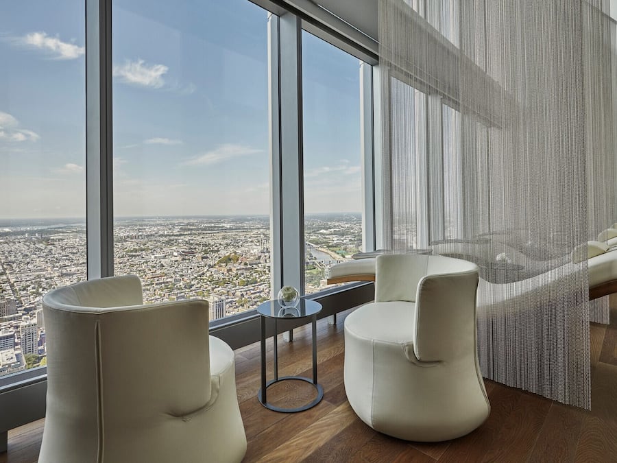Una habitación moderna con un ventanal amplio que ofrece vistas a la ciudad, dos sillas blancas, una mesita pequeña y una cortina decorativa.