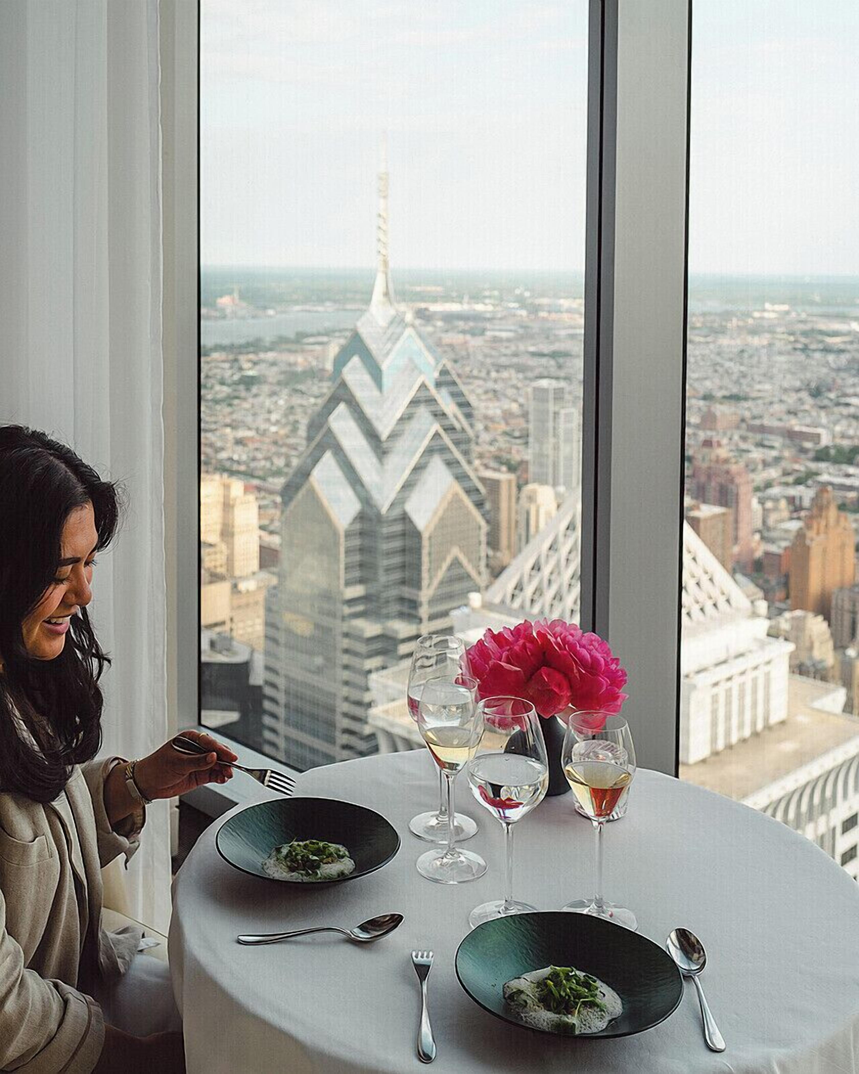 Een eettafel met borden eten, wijnglazen en een bloevas, met op de achtergrond een stadsgezicht vanuit een hoog gebouw.