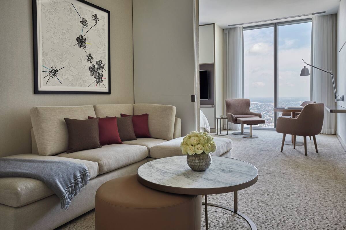 Ein modernes Hotelzimmer mit einer Couch, einem runden Couchtisch mit Blumenvase, einem Essbereich mit Stühlen und einem großen Fenster mit Blick auf die Stadt.