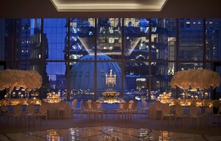 Comedor formal con mesas dispuestas para seis personas, ventanas amplias con vista a la ciudad y arreglos florales blancos a cada lado.