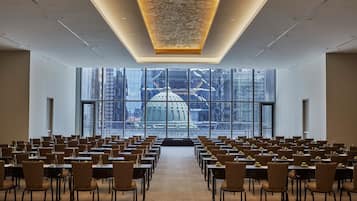 A conference room with rows of tables and chairs, large windows with a view of a cityscape, and a modern ceiling design.
