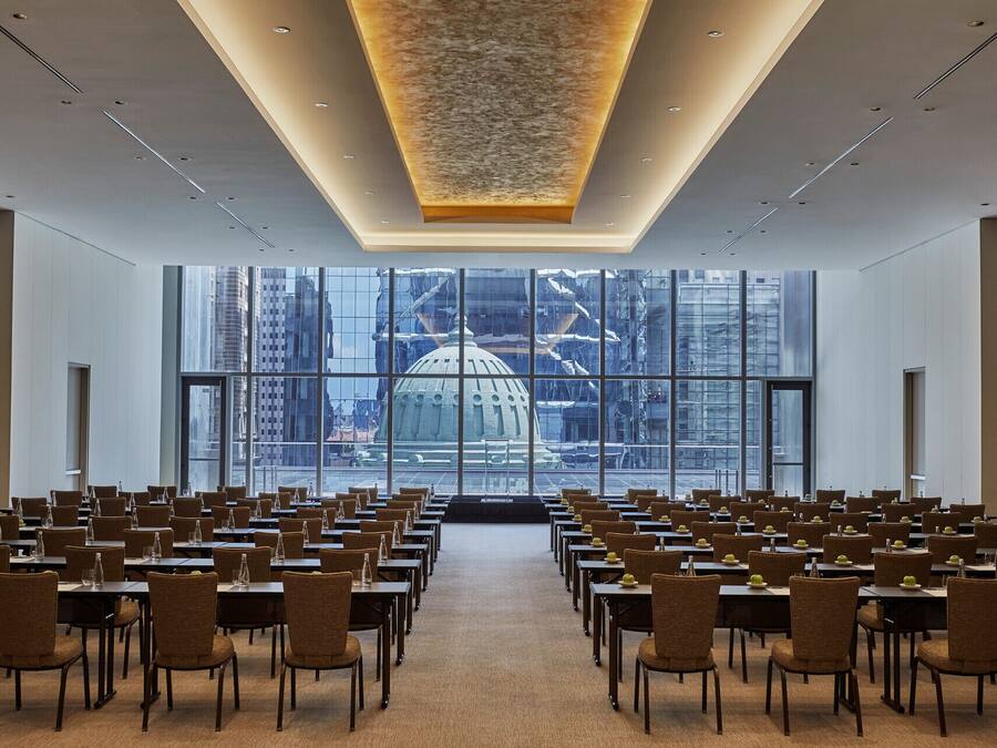 A conference room with rows of tables and chairs, large windows with a view of a cityscape, and a modern ceiling design.