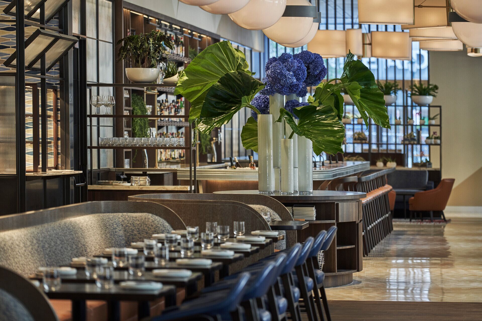 A modern restaurant with a long counter, multiple tables, and a variety of plants and decorative items.