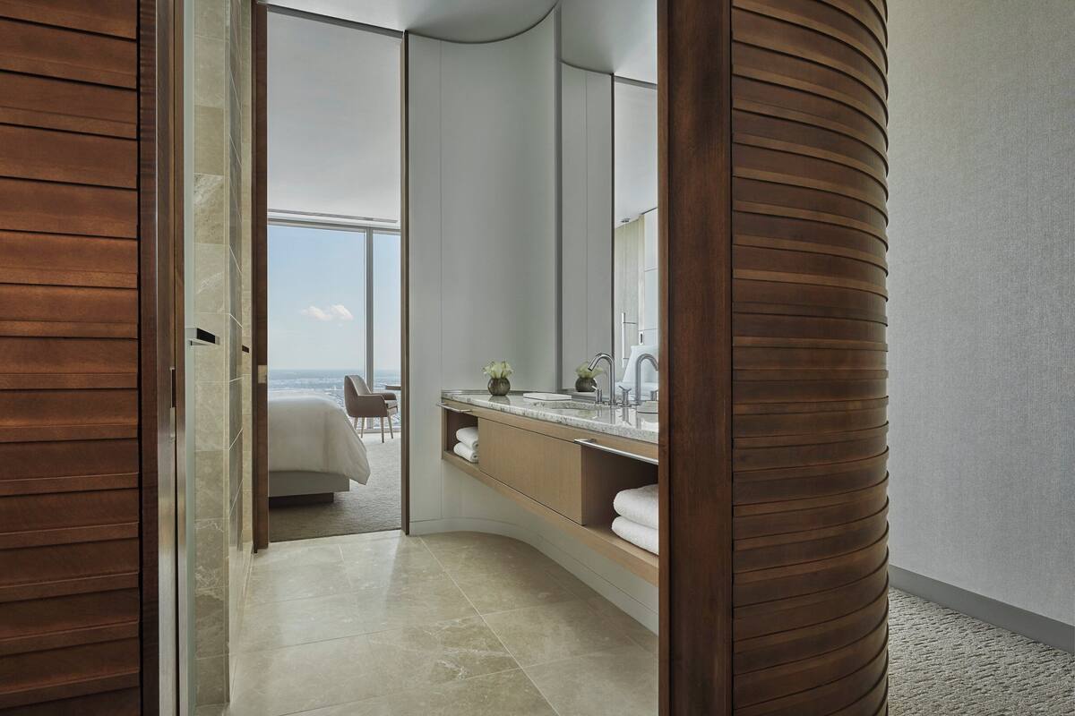 Ein modernes Badezimmer mit großem Spiegel, Waschbecken und Blick auf ein Schlafzimmer mit Bett und Sessel.
