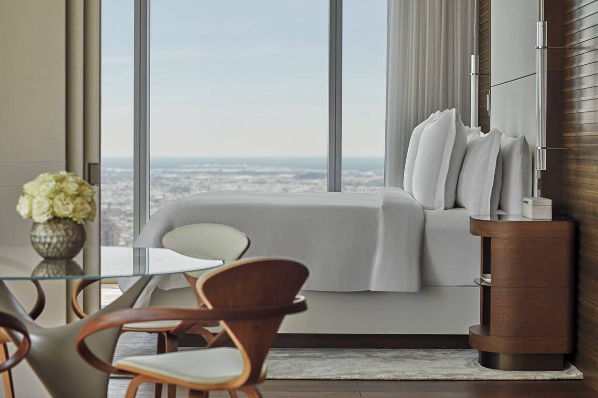 Ein modernes Hotelzimmer mit großem Fenster und Blick auf die Stadt, einem Bett mit weißer Bettwäsche, einem hölzernen Nachttisch und einem Glastisch mit Stühlen.