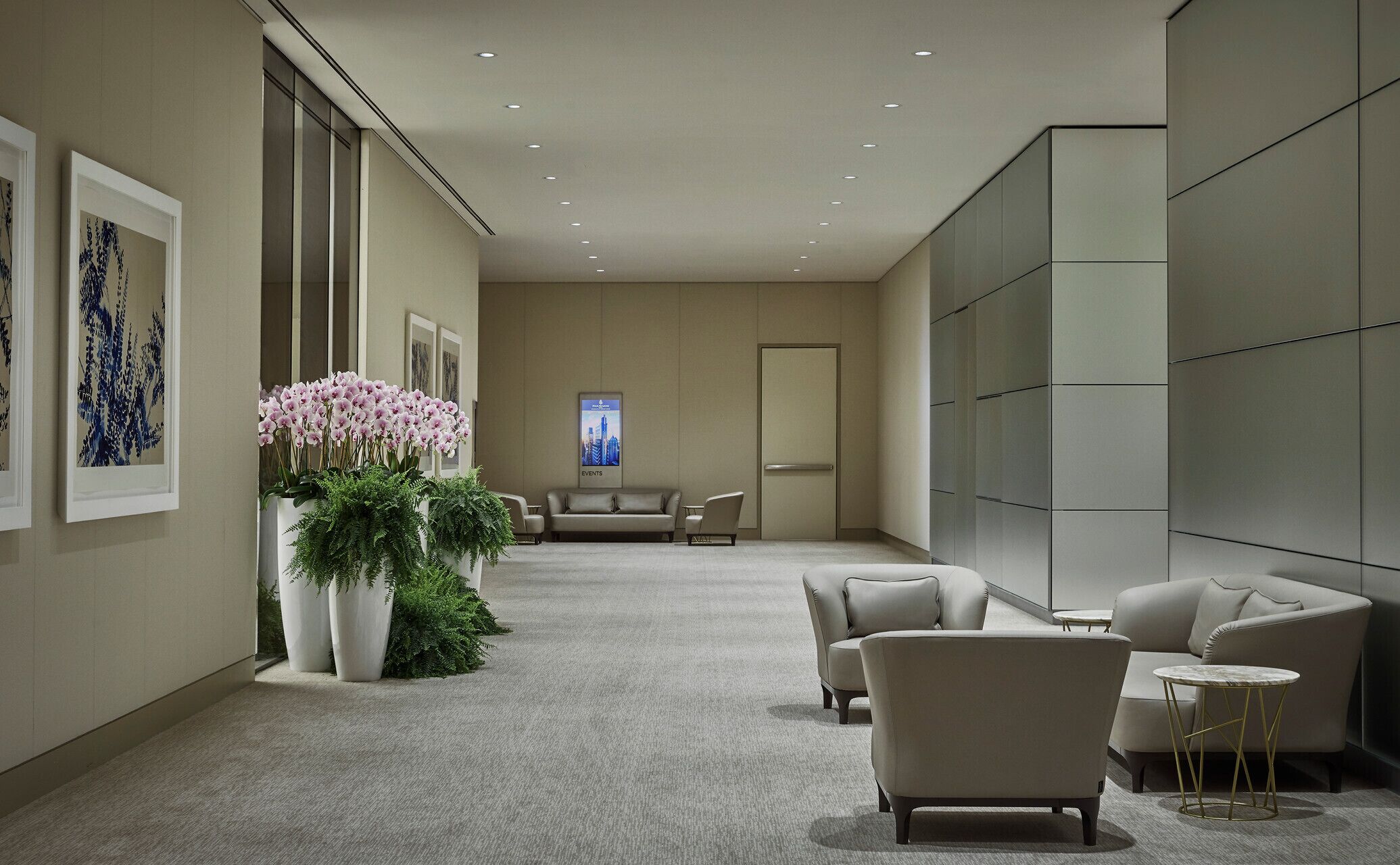 A modern hotel lobby with a neutral color palette, featuring a seating area with sofas and armchairs, a large potted plant with pink flowers, and framed artwork on the walls.