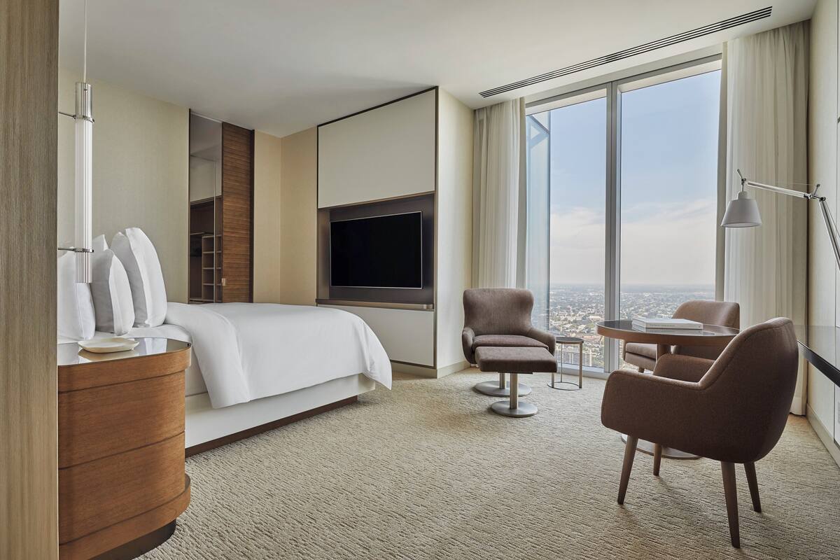 Ein modernes Hotelzimmer mit einem großen Bett, einem Flachbildfernseher, einer Sitzecke mit Tisch und Stühlen und Blick auf die Stadt durch große Fenster.