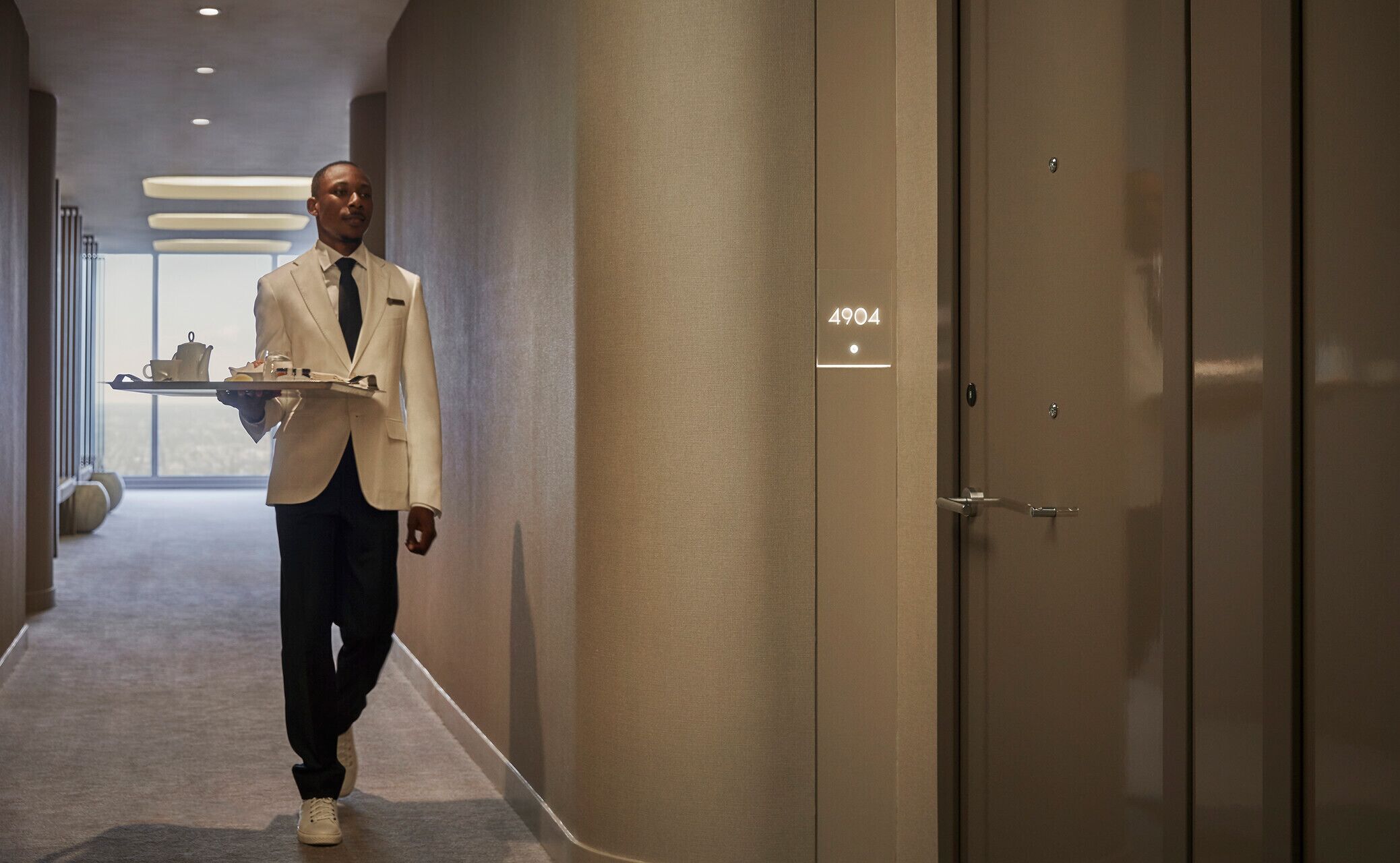 A hotel corridor with a person in a white suit carrying a tray with a teapot and cups. The corridor has a number '4904' on the wall and a door with a handle.