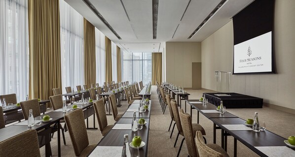 Un salón de conferencias con mesas largas, sillas y una pantalla de proyección que muestra el nombre del hotel.