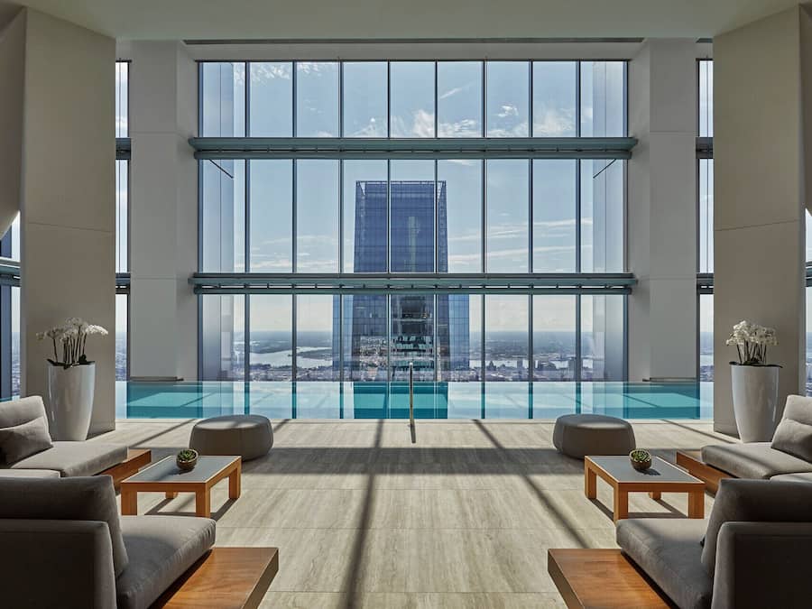 Un salon moderne doté d’une piscine à débordement en verre, de grandes fenêtres et d’une vue sur la ville.